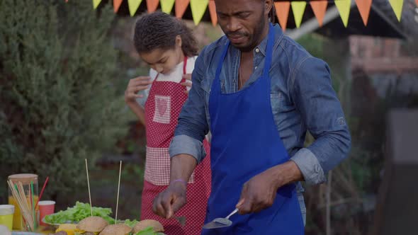 African American Focused Man Cleaning Spatula with Fork Standing on Picnic at Barbecue Grill alt