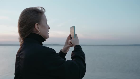 A Woman Takes Pictures on Her Phone of a Sunset on a Lake on an Autumn Evening