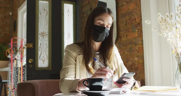 Caucasian female customer wearing face mask, sitting at table, using smartphone alt