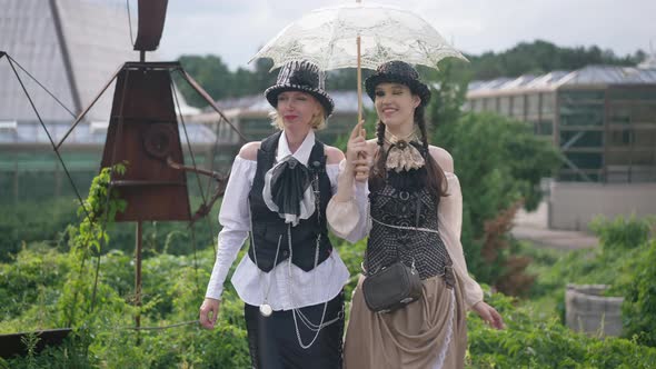 Cheerful Beautiful Mother and Daughter in Steampunk Costumes Strolling with Sun Umbrella Leaving alt