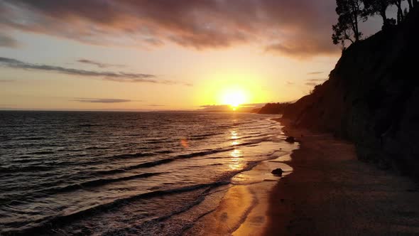 Aerial drone shot of an orange sunset sky above the ocean waves off the sandy beach cliffs of Santa alt