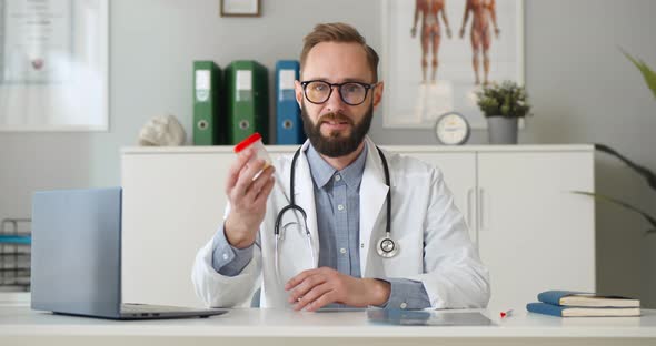 Male Doctor Talking to Camera Holding Bottle with Pills During Telehealth Mobile Consultation alt