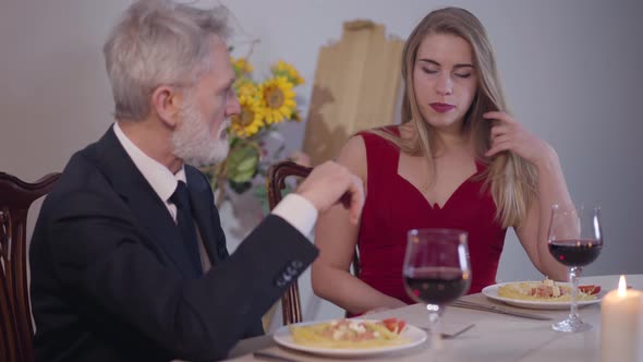 Portrait of Young Seductive Woman in Red Dress Sitting with Senior Man at the Table and Smiling alt