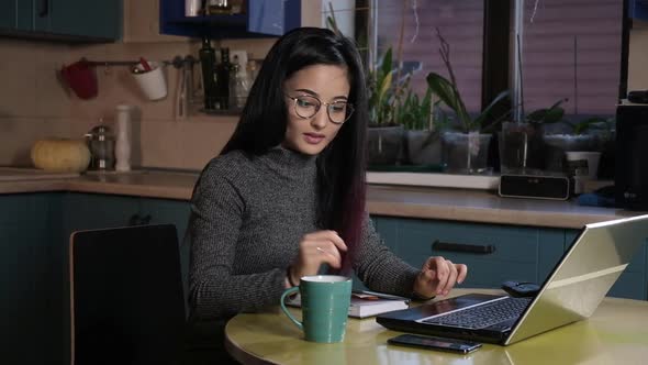 Asian woman in evening sitting in kitchen at home working on a laptop. alt