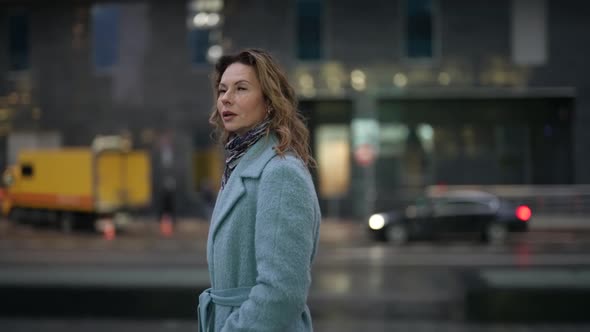 a Blonde in a Light Coat Has Hands in Pockets Looks at the Camera Smiles and Walks in the Twilight alt