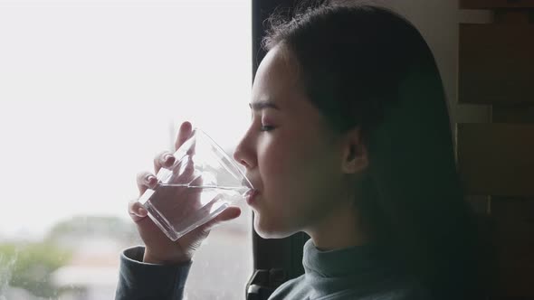 Beautiful Asian female drinking water while standing beside a window in the kitchen at home.