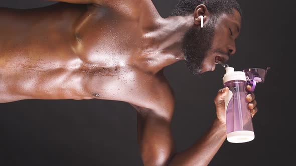 An AfricanAmerican Man with Bare Breasts on an Isolated Dark Background Drinks Water After a Workout