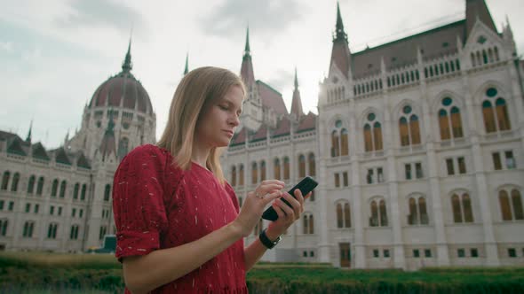 Woman in Red Dress Uses Mobile App on Smartphone By Budapest Parliament Building