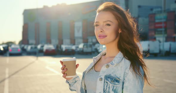 A Woman is Standing with a Cup of Coffee in the Parking Lot of a Shopping Center alt