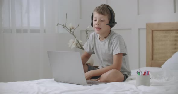 Cheerful Boy Is Communicating with Someone By Video Call on Laptop, Using Headphones, Sitting on Bed alt