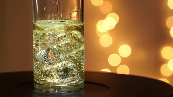 Champagne is Poured Into Glass with Ice and Rotates on Tray on Background of Luminous Golden Bokeh