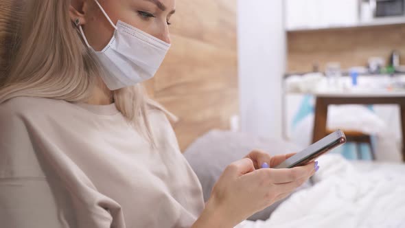 Sick Beautiful Woman in Medical Mask Lying in Bed with Smartphone