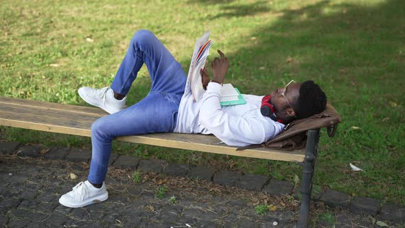 Wide Shot Relaxed Confident African American Student Lying on Bench Outdoors Learning Material alt