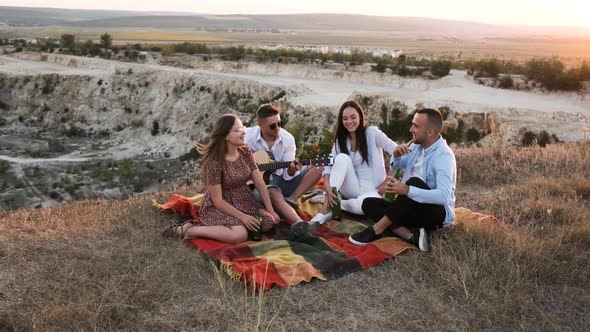 Four Friends Cheering with Beer and Smiling While Sitting on the Blanket at Picnic alt