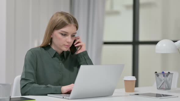 Young Woman Talking on Smartphone While Using Laptop alt