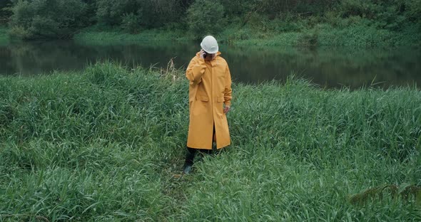 Worker in Helmet and Raincoat Walks to River on Grass Man Has Phone in His Hand alt