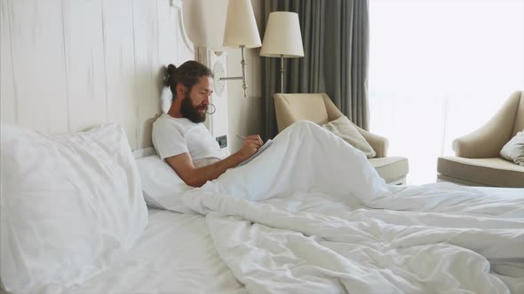 Young Bearded Man Writing Notes to Diary After Awake in Bedroom at Home