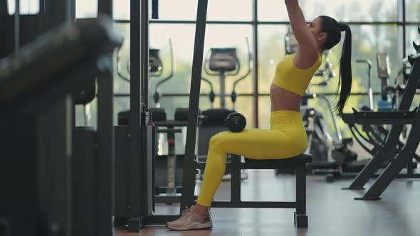 Hispanic Woman Sitting on a Simulator in the Gym Pulls a Metal Rope with the Weight Pumps Up the alt