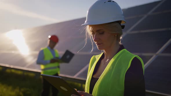 Portrait of a Female Solar Engineer Standing Uses a Tablet and Looking at the Camera Another Man