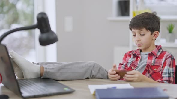 Relaxed Middle Eastern Little Boy Sitting with Feet on Table and Messaging Online on Smartphone alt