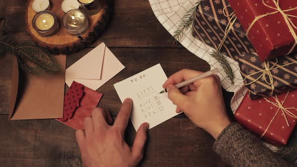 Top View of Mans Hands Putting Ticks in the Gifts List for His Family on Wooden Christmas Decorated alt