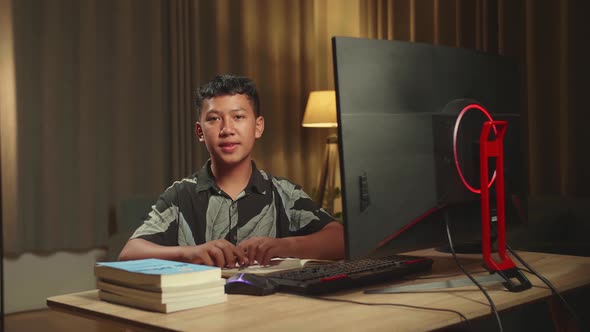 Asian Boy Learning Online With Desktop Computer From Home, He Turns And Smiles Into The Camera alt