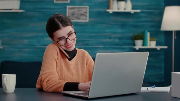 Young Woman Talking on Phone Call and Using Laptop at Home Desk alt