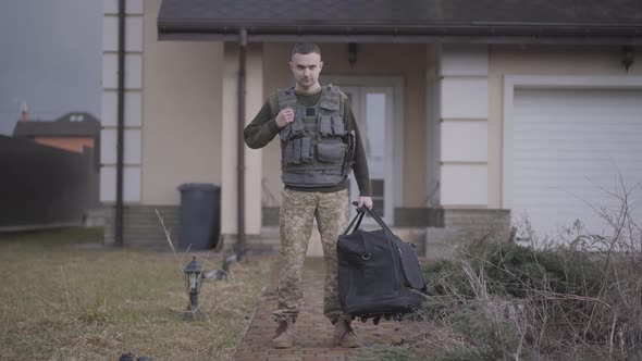 The Young Soldier in Military Closes with Big Bag Standing in Front of the House alt