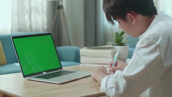 Asian Man Student Look At Green Screen Computer And Write In The Notebook While Studying Online alt
