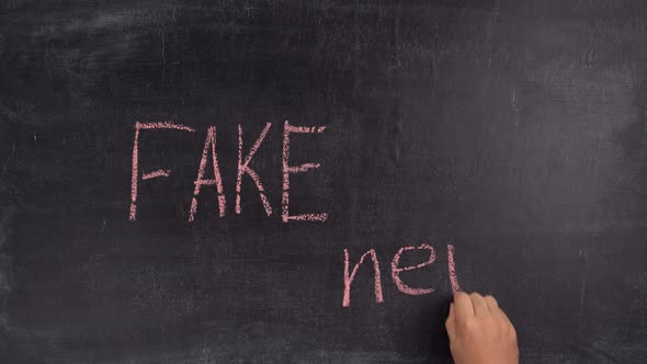 A man's hand writes the phrase FAKE NEWS in red chalk on a black chalk board alt