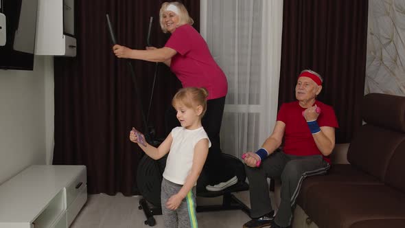 Senior Couple Using Orbitrek Doing Dumbbells Exercises Child Girl Making Selfie on Mobile Phone alt
