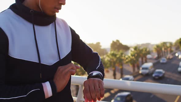 Midsection of african american man wearing earphones exercising on footbridge checking smartwatch alt