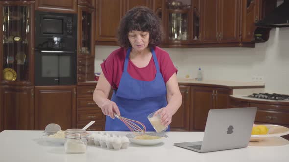 Senior Caucasian Woman Checking Recipe Online and Mixing Milk Into Dough alt