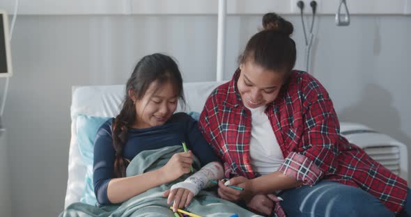 Smiling Chinese Teen Girl Lying in Hospital Bed and Painting Hand Gypsum with Foster Mother alt