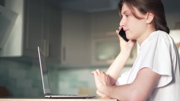 The Woman Works Behind a Laptop and Speaks on the Phone in Her Kitchen. A Successful Freelancer on