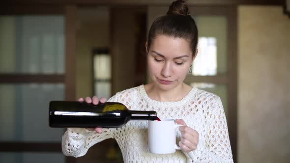 Girl pours red wine into a mug and drinks, looking out the window in the kitchen alt