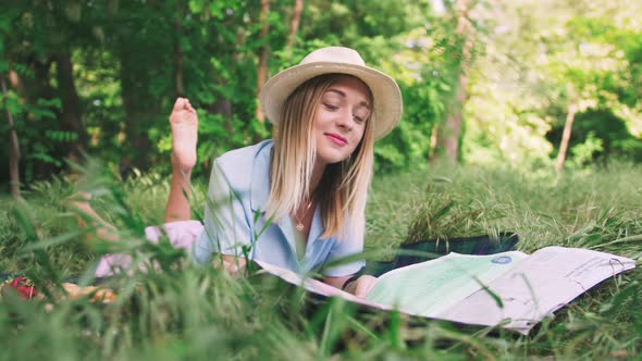 Portrait of Happy Romantic Young Woman in Hat Reading Magazine Outdoors in Park on Picnic alt
