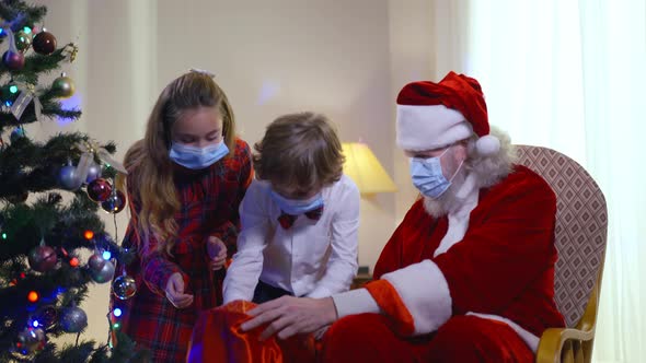 Happy Children Taking Gift Boxed From Santa Bag As Senior Bearded Man Sitting in Armchair in Living alt