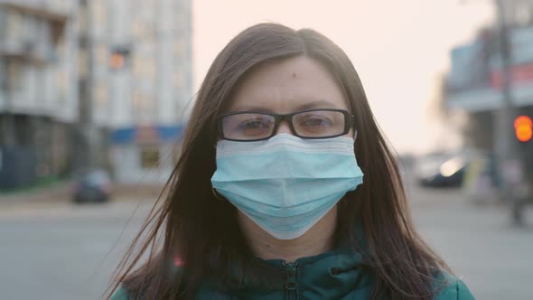 A Young Girl in Glasses and a Medical Mask Stands on the Street alt