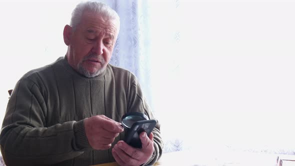 Old grandpa uses a magnifying glass to read from his smartphone. alt