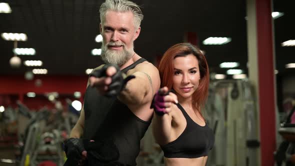 A sporty man with a beard and a young woman point a finger at the camera alt