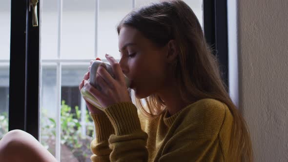 Woman drinking coffee while looking out of the window at home alt