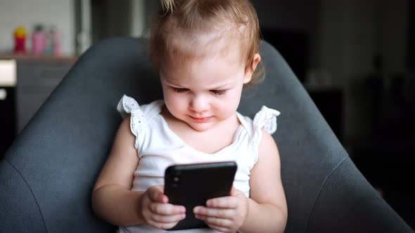 Smiling Little Girl is Sitting on Armchair and Using Smart Phone alt
