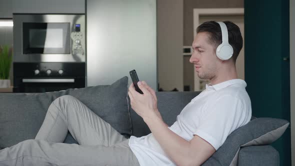 Portrait of Cheerful Young Man Lying on the Sofa and Using Smartphone Application Listening to Music alt