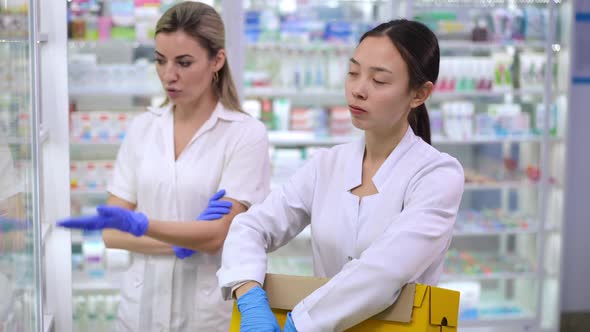 Overburdened Asian Young Pharmacy Assistant with Box Standing at Shelves with Angry Caucasian alt