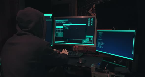 Hacker in a Hood Sits in Front of Computer Screens and Hacks Databases