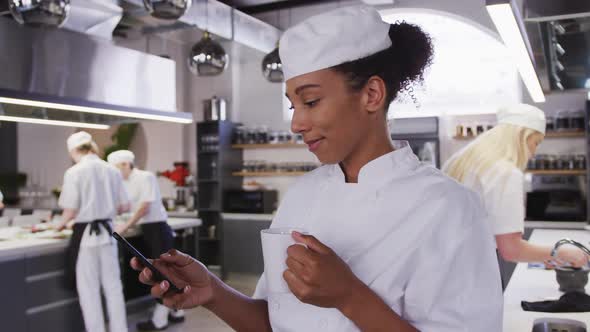 African American female chef wearing chefs whites in a restaurant kitchen using a phone and smiling alt