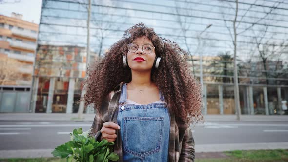 Beautiful African American Woman in Eyeglasses and Headphones is Going Home From the Market with alt