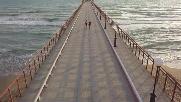 Aerial view of teenagers boy and girl walk by hand on pier in sea foaming waves love on beach summer