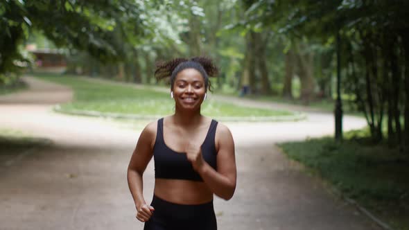 Follow Shot of Sporty African American Woman Jogger Running in Public Park Listening to Music and alt